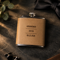 Personalized groomsmen flask in tan leather, engraved GROOMSMAN, JUSTIN and the wedding date, resting on dark slate with a black bow tie, cufflinks and eucalyptus