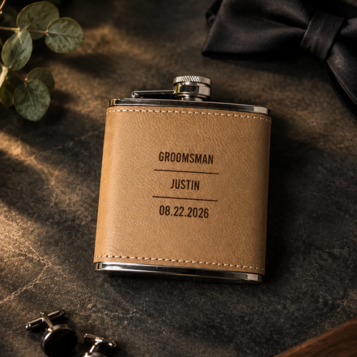 Personalized groomsmen flask in tan leather, engraved GROOMSMAN, JUSTIN and the wedding date, resting on dark slate with a black bow tie, cufflinks and eucalyptus