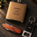 The Signature Set for groomsmen: an engraved leather flask, a rosewood pocket knife and a leather keychain bottle opener together on dark slate, each engraved with his name
