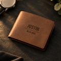 Personalized groomsmen bifold wallet in brown leather, engraved JUSTIN above the wedding date, on dark slate beside a wristwatch and eucalyptus